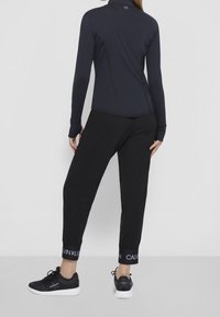 Osoba v černých teplácích s logem Calvin Klein na manžetách a černých teniskách, stojící zády k fotoaparátu na šedé podlaze.