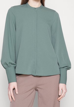 Femme portant un chemisier vert sourd à manches longues avec un col boutonné et un pantalon large taupe sur un fond uni.