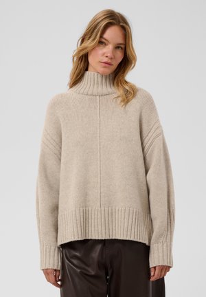 Femme aux cheveux blonds mi-longs, portant un pull à col roulé beige et un pantalon marron foncé, debout devant un fond clair uni.