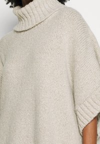 Flauschiger Strickpullover in hellem Beige mit umgeschlagenem Rollkragen, kurzen Ärmeln und einem leicht offenen Strickmuster, das dezente Glanzakzente setzt.