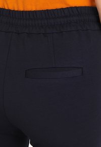 Marinblå byxor med en slät textur, utrustade med ett elastiskt midjeband och en liten bakficka med en rektangulär klaff.