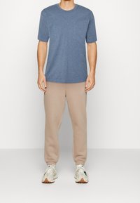 Mand iført en blå t-shirt med kortærmer, beige joggebukser og hvide sneakers, der står foran en ensfarvet lys baggrund.
