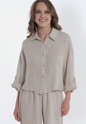 Donna sorridente con capelli ricci di media lunghezza, che indossa una camicia beige di lino con bottoni, maniche arrotolate e pantaloni larghi abbinati.