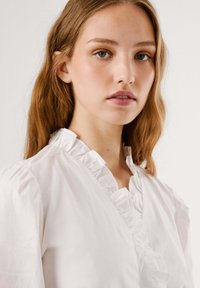 Giovane donna con capelli castano chiaro mossi che indossa una camicia bianca con colletto a balze su uno sfondo chiaro e uniforme.