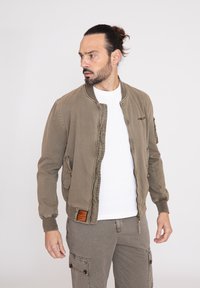 Chaqueta bomber verde oliva con cierre de cremallera, bolsillos laterales y un parche en el dobladillo. Usada sobre una camisa blanca y combinada con pantalones claros.