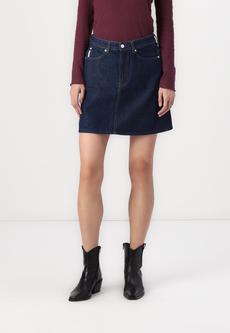 Denim mini krilo, temno modro, s prilegajočim pasom, sprednjimi žepi in vidnimi šivi. V kombinaciji z črnimi gležnjarji in rebrastim maroon topom.