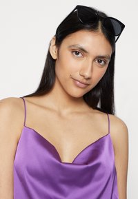 Camisola de cetim roxa com alças finas, apresentando um design de decote em V profundo. Um par de óculos de sol estilo gato preto repousa sobre cabelos longos e escuros.