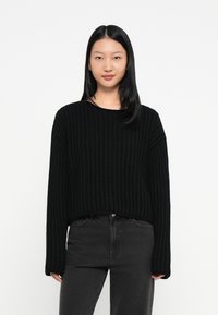 Giovane donna con capelli neri lisci che indossa un maglione a maglia nera testurizzata e jeans scuri a vita alta, in piedi contro uno sfondo bianco.