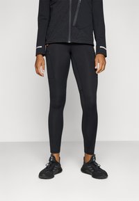 Tenue de sport noire composée de leggings ajustés avec une poche latérale, accompagnée d'une jacket noire ornée de motifs géométriques discrets et d'accents réfléchissants.