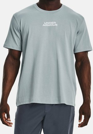 Man wearing light blue Under Armour short-sleeve T-shirt and dark shorts, standing against a plain white background.