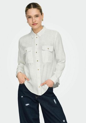 Kobieta z włosami związanymi do tyłu, nosząca złote kolczyki-koła, białą koszulę zapinaną na guziki z kieszeniami oraz ciemne, przetarte dżinsy, trzymająca ręce w kieszeniach.