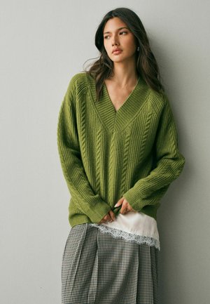 Junge Frau mit langen Haaren, die einen grünen Kabelstrickpullover über einem weißen, mit Spitze verzierten Oberteil und einem karierten Rock trägt, steht vor einer schlichten Wand.