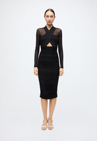 Black ruched dress with long sheer sleeves, a cut-out at the waist, and a fitted silhouette. Model wears silver strappy heels.