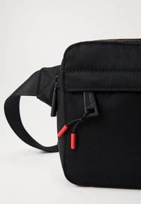 Borsa in tessuto nera a vita con cintura regolabile, tasca frontale con zip e linguette rosse sulle zip.