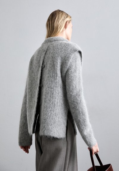 Szürke, textúrált kardigán, laza fazonnal, V-nyakkivágással, nagy gombokkal a háton és hosszú ujjal. Elegáns nadrágra van húzva.