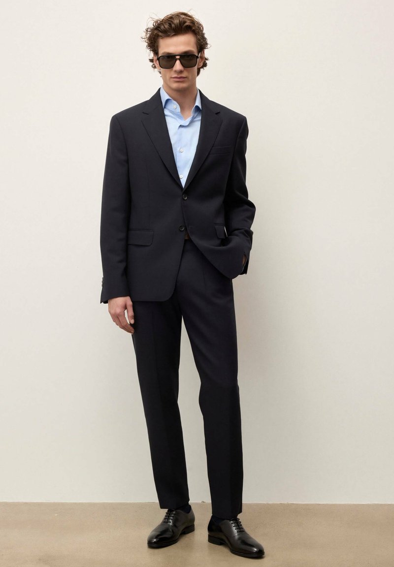 Muž oblečený v čiernom obleku s bledomodrou košeľou, čiernymi spoločenskými topánkami a tmavými slnečnými okuliarmi, stojí pred jednoduchým svetlým pozadím.