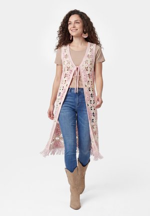Kvinde med krøllet hår iført en pink hæklelang vest over en beige t-shirt, blå jeans og brune cowboystøvler, smilende og gående fremad.