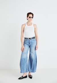Débardeur blanc à côtes, jeans bleus clairs à taille haute et jambes larges légèrement délavés, ceinture noire cloutée et chaussures plates noires. Lunettes de soleil rondes.