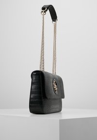 Černá kabelka z texturované kůže s klopou, zlatými kovovými doplňky a páskem s dvojitým řetízkem. Na přední straně má výrazné logo.
