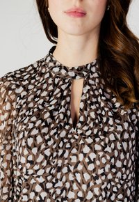 Blusa in stampa leopardata marrone e bianca con scollatura a chiave, dettaglio plissettato e texture trasparente. Presenta un colletto alto e maniche fluide.