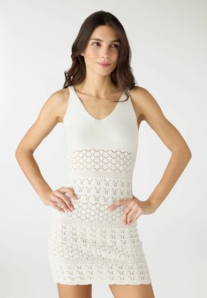 Donna con capelli lunghi e scuri che indossa un abito bianco senza maniche in maglia con lavorazione traforata a motivo, mani sui fianchi, che guarda leggermente di lato.