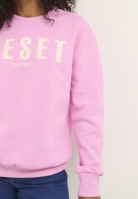 Persona con un sudadera rosa que dice "RESET RECHARGE" impresa en la parte delantera, combinada con jeans azules, frente a un fondo blanco.
