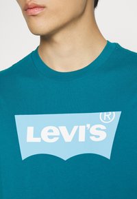 T-shirt en coton teal arborant un grand logo Levi's bleu clair en forme de courbe, avec un col rond et une coupe standard.