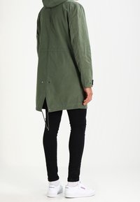 Manteau long vert avec capuche, fabriqué à partir d'un tissu lisse. Comprend une ventilation arrière, des réglages ajustables et des fermetures à pression. Porté par-dessus un pantalon noir.