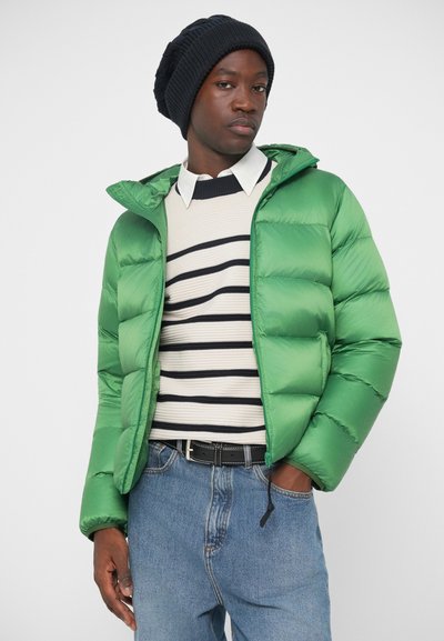 Zöld puffermellény kapucnival, könnyű anyagból, steppelt részekkel, cs�íkos pulóver fölött viselve, kék farmernadrággal és fekete övvel párosítva.