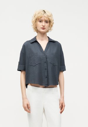 Νέος με κοντά, σγουρά ξανθά μαλλιά, που φοράει κοντό γκρι σκουρό πουκάμισο με κουμπιά και λευκό παντελόνι, στέκεται μπροστά σε απλό φόντο.