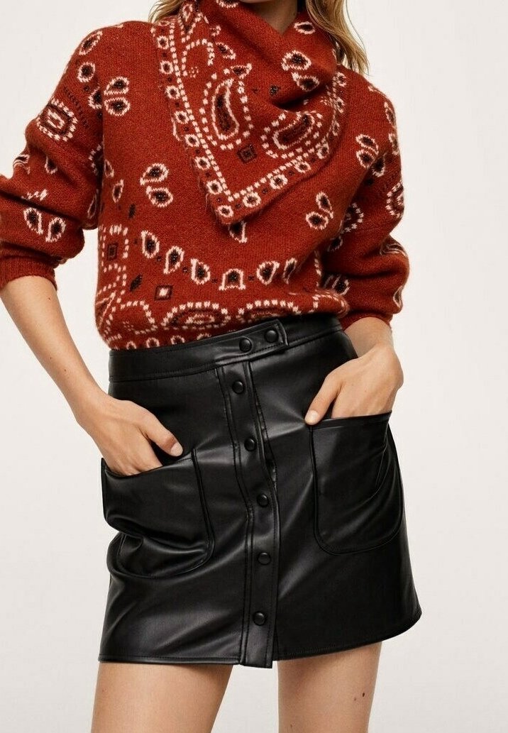 Femme portant un pull à motifs rouge rouille rentré dans une mini-jupe en cuir noire à boutons avec de grandes poches devant et les mains dans les poches.