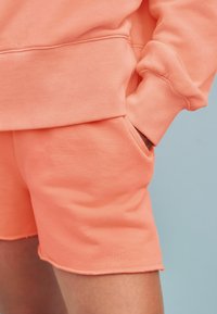Koral sweatshirt med ribbet manchetter, parret med matchende koral shorts med en lomme og rå kant. Blød tekstur og afslappet pasform.