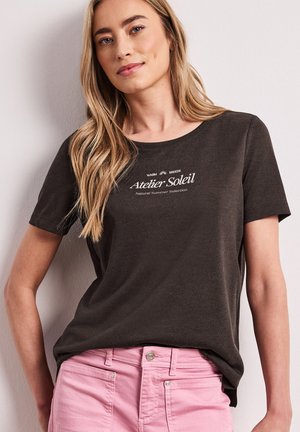 Femme blonde portant un tee-shirt marron foncé avec le texte « Atelier Soleil » et un pantalon rose clair, debout contre un mur clair.