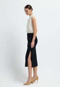 Cremfarbener ärmelloser Top mit strukturiertem Design, kombiniert mit einem schwarzen rockartigen Midiskirt mit seitlichem Schlitz. Das Model trägt beigefarbene, geflochtene Absätze.