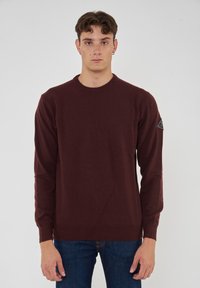 Maglione lavorato a maglia bordeaux con maniche lunghe, scollatura rotonda e polsini a coste. Presenta una finitura testurizzata e un patch con logo sulla spalla sinistra.