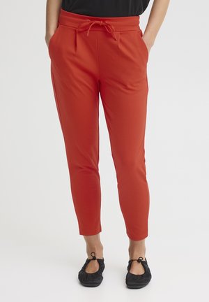 Woman wearing slim-fit burnt orange pants with drawstring waist and black slip-on shoes, standing with hands in pockets.