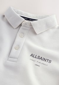 Bílá polokošile s límečkem a dvěma knoflíky, na prsou s logem "ALLSAINTS" a japonským textem nad nápisem "LONDON".