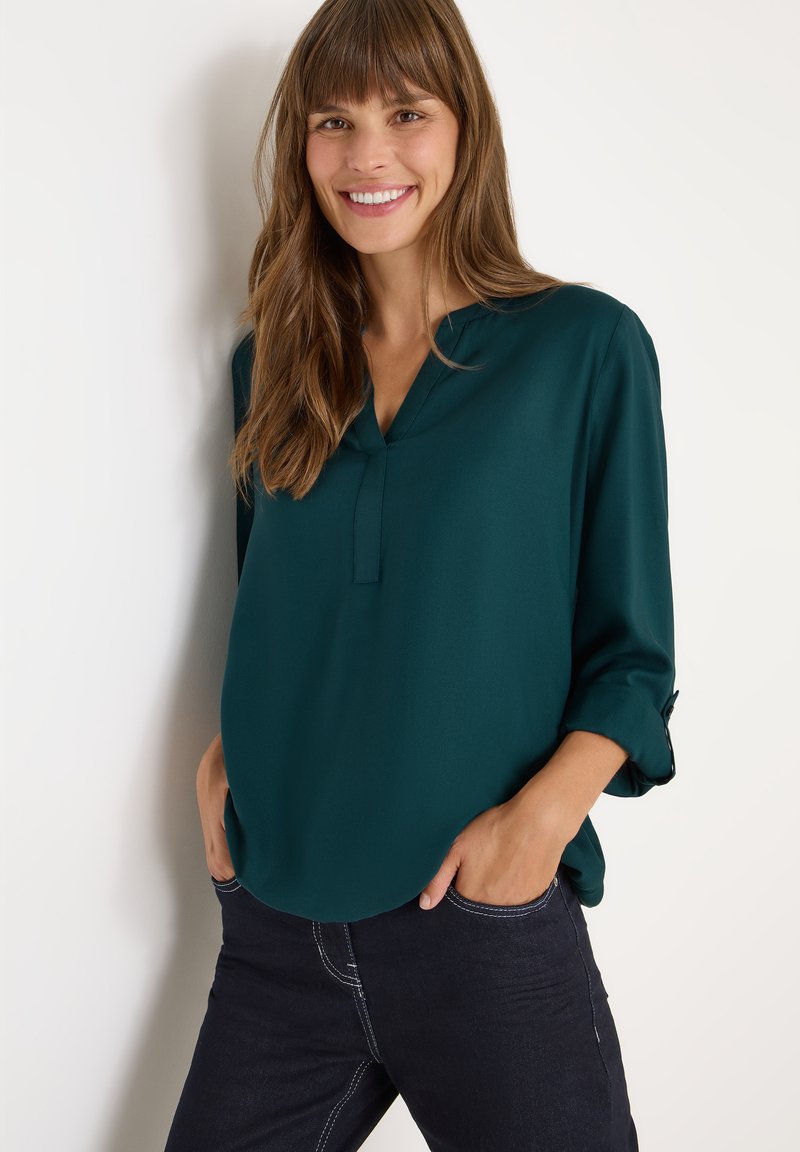 Blouse teal en tissu lisse, avec un décolleté en V et des manches retroussées, associée à un jean foncé avec des coutures contrastantes.