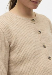 Bež pletena kardigan z valovitim vzorcem, okroglim izrezom in petimi rjavimi gumbi. Tekstura se zdi mehka in rahlo puhasta.