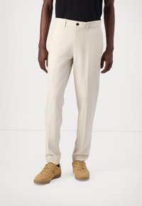 Beige kalhoty s rovnou siluetou, vyrobené z lehkého materiálu, s předním zapínáním na knoflík a bočními kapsami. Nosí se s béžovými teniskami.