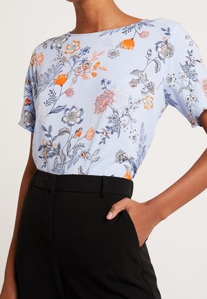 Lyseblå bluse med blomsterprint i orange og sort. Korte ærmer, rund halsudskæring, parret med højtaljede sorte bukser. Glat stof.