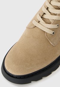 Bota de ante beige con punta redonda, que presenta cordones del mismo tono y una suela de goma negra. Textura suave con detalles de costura bien acabados.