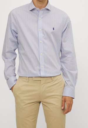 Hombre con camisa de rayas verticales azules y blancas abotonada y pantalones beige, de pie con las manos en los bolsillos.