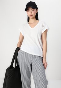 Vit V-ringad t-shirt med korta ärmar, matchad med randiga ljusgrå och vita byxor. Modellen bär en svart tote-väska.