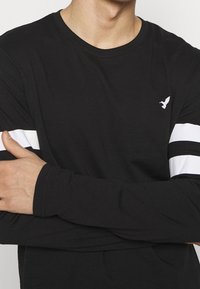 Svart långärmad tröja i bomull. Har vita horisontella ränder på ärmarna och en liten vit örnlogotyp på bröstet.