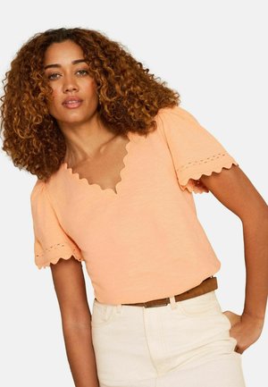 Žena s kučeravými vlasmi, oblečená v broskyňovom tričku s krátkym rukávom s priečnym lemom a v bielych vysokých nohaviciach s hnedým opaskom.