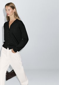 Black button-up cardigan with a v-neckline, long sleeves, and ribbed hem, paired with white wide-leg pants. Simple, textured fabric.
