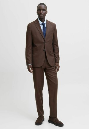 Brown suit with a single-breasted jacket, paired with a white shirt and navy tie. Worn with dark brown textured shoes. Plain background.