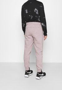 Persoon die lichtroze joggingbroek, een zwart tie-dye sweatshirt en zwart-witte Nike sneakers draagt, staand tegen een effen achtergrond.
