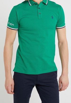 Mann trägt grünes kurzärmliges Poloshirt mit weißem und marineblau gestreiftem Kragen und Ärmelsaum, kombiniert mit dunkelblauer Hose.